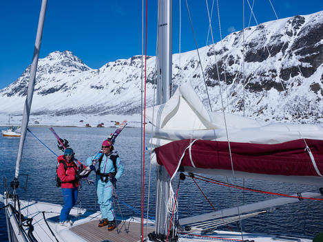 Two skiers on moored sailing yacht with fjord and snowy mountain in the background