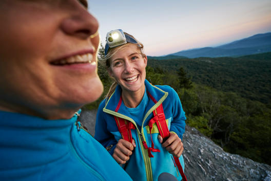 An older woman and a younger woman laughing while adjusting their headlamps on a scenic rocky outcropping at dusk while on a backpacking trip together.