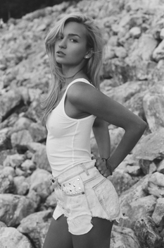 Black and white photo of blonde woman in cut-off jeans and white tank top slightly arched taken from the side with rock background