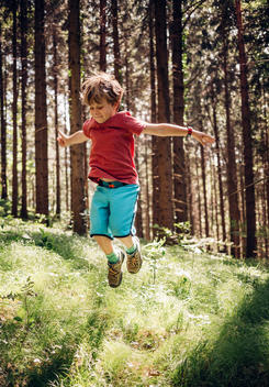 6 year old blonde boy in blue shorts and red shirt jumping in gras in forest in Austria.