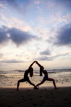 Two female yoga practitioners on the beach at sunrise back to back doing a warrior one pose, reaching backwards in a silhouette shot