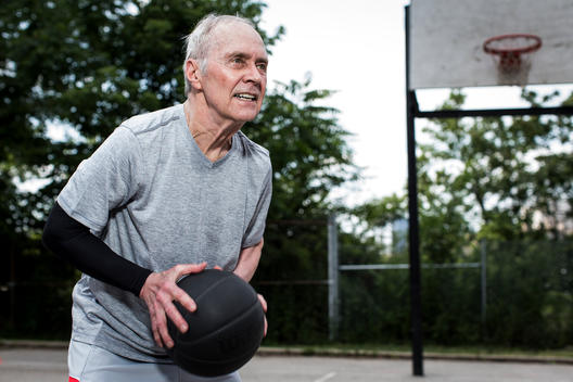 Portrait of an older 50-plus man playing basketball outside on an asphalt court in an urban landscape.