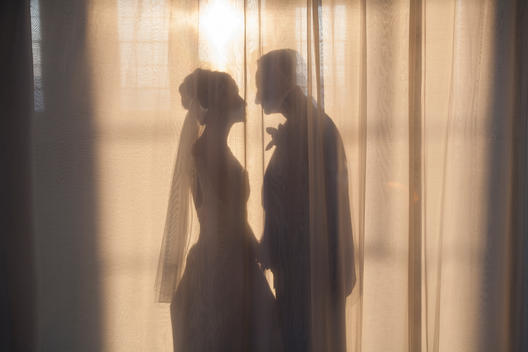 Silhouette of bride and groom facing each other behind sheer curtain