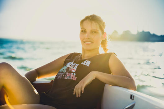 young woman wearing a fashionable outfit while sitting on a boat at summer and facing in camera, back light and lens flare