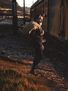 cool beaded man walking by a train track with a sack on his back