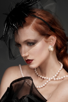 Studio beauty cropped portrait red haired female model in a veil, pearls ad a black dress, simple styling and a long ponytail she looks off camera in front of a dark background