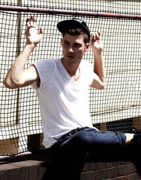 Young Male In A Base Ball Cap, White T Shirt And Jeans Sitting On A Brick Wall
