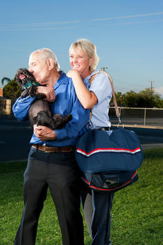 A female mail carrier hugs the back of a man holding a dog