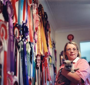 Portrait Of A Woman Holding Her Burmese Cat Standing Next To A Wall Covered With Ribbons