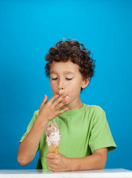 A young boy in a green shirt licks his finger as he holds an ice cream cone in the other hand