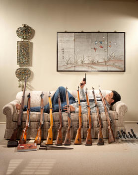 Man is laying on couch holding a gun with many guns leaning against him