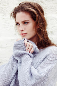 closeup of brown haired model in blue sweater holding her hands under her chin looking away from the camera