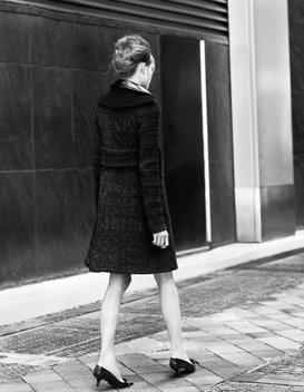 A female model walks along the sidewalk outside a city building with a view from the back.