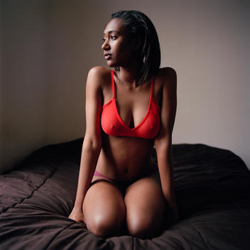 Portrait of young tall African-American black teenage girl wearing a red lace bra and purple panties lingerie with hard erect nipples squatting on a bed and gazing out of the window. Brooklyn, New York