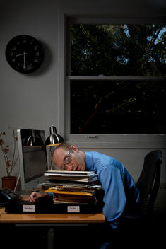 A balding man in a blue dress shirt sleeps on a pile of paperwork on his desk