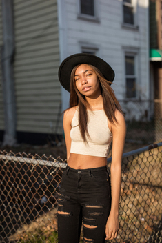 Portrait of cute African-American/black teen girl and model in crop top leans against a chanlink fence on a sunny afternoon during a street fashion shoot. Bed-Stuy, Brooklyn