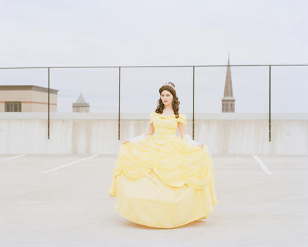 Portrait of Sapphire Nova, an ordinary woman who works as and impersonates Disney Princess Belle in yellow dress standing on the top floor of an empty parking garage in downtown with a church steeple behind. New Jersey