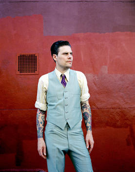 Musician Matty Charles Wearing A Suit And Showing Tattooed Arms Standing Against A Red Wall.
