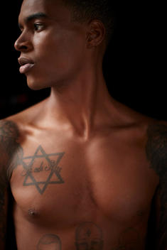 Close-up of shirtless man with tattoos on black background.