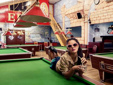 Young model in gold top and sunglasses lying on edge of pool table in eclectic wall painted pool club.