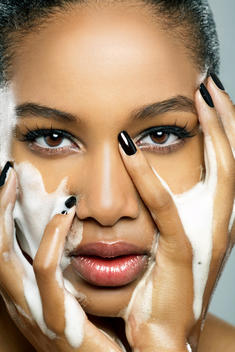 African American model beauty close up of her face, looking at you holding her face with her hands wearing black nail polish and having foam on her face and her hands washing her face