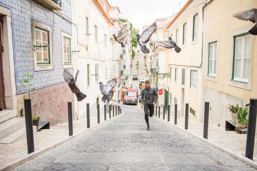A man running up the center of a street and breaking up a flock of pigeons as they fly around him with tall colorful buildings stretching back in the alleyway behind him in the city of Lisbon, Portugal