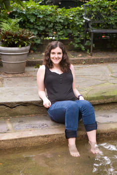Young Woman Sitting By The Garden Fountain Comfortably Dipping Her Feet In The Water