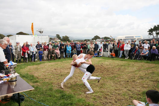 Crowds gather at Gosforth Agricultural Show to watch two boys compete in a Cumberland and Westmorland wrestling match. The boys are dressed in traditional Cumberland wrestling costumes.