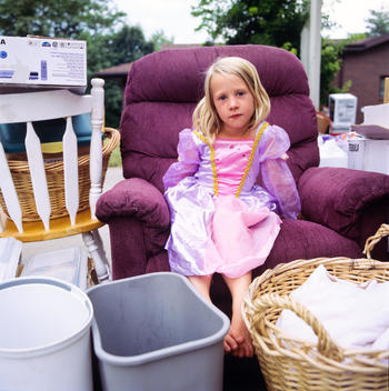 Girl Sits On Chair Amidst Other Household Stuff Strewn On Front Lawn
