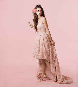 Brunette woman in coral pink dress and gold heels on pink studio background with flower in hair