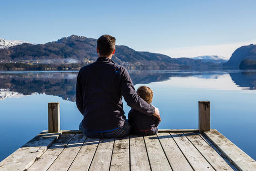 Father and son sitting by a calm Fjord with snow covered mountains in the distance, Myking, Norhordaland, Norway.