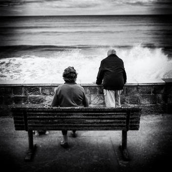 Elderly couple (woman seated on bench) and man (looking over wall) watch the high waves crashing in. Dog also present, just visable under bench.