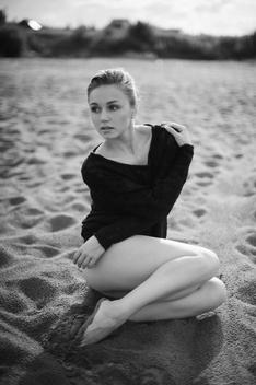 blonde girl in a black bodysuit and black sweater sits gracefully on the sand.