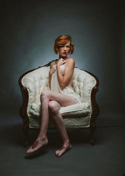 Nude woman, redhead covered only in sheer fabric. Sitting on white chair, neutral background