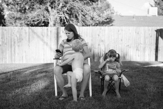 A pregnant young mother busy on her cell phone ignores her young son in diapers as he tries to get her attention and daughter eats a bowl of cereal in the backyard on a sunny summer afternoon. Denver, Colorado