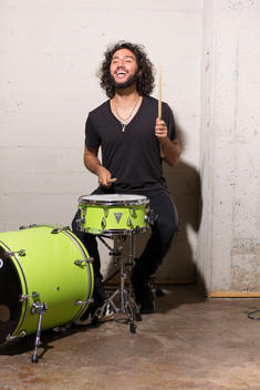 Smiling mixed ethnicity millennial man in loft playing green drum set hair flying.