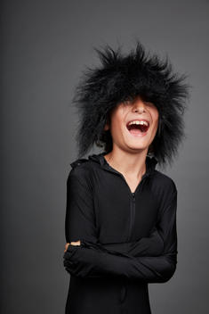 A boy wearing a wig that covers his eyes laughs with his arms crossed in front of him