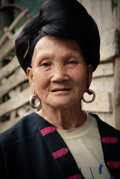 Portrait Of A Miao (Ethnic Group) Old Woman In Traditional Clothes And Earrings Smiling. Women In This Area Have Long Hair Gathered In A Knot On The Top Of The Head.