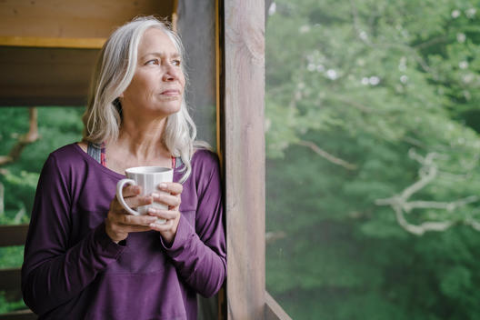 Fit older woman with silver gray hair drinking coffee in a screened in room wearing yoga clothes.