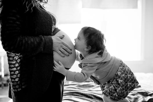 Black and white image of pregnant mom and young daughter kissing belly