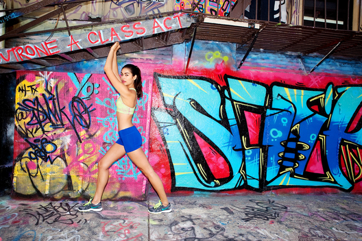 Location outdoor in front of graffiti walls caucasian/Spanish female fitness model stretching in athletic clothes looking into camera