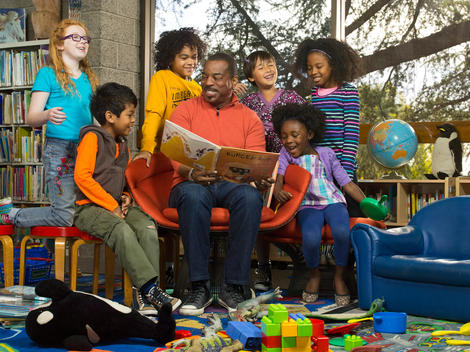A group of children listen to Actor LeVar Burton read them a book in a library