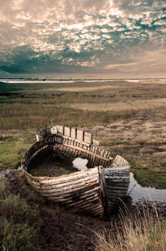 Decaying timber fishing boat resting on marshland in water at Blakeney in North Norfolk, England