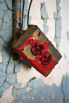 Close-Up Of Ac Electric Outlet Box With Peeling Red Paint On Chipped-Paint Wall