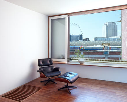 Clean white room with wooden floor and large view through window overlooking London Waterloo and the millennium wheel. Black leather Eames chair and foot stool sit on floor.