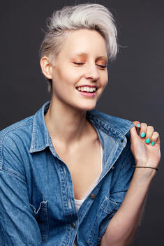 laughing fashion model in studio with closed eyes, in short blond hair, orange eye makeup and light pink lip wearing a jeans shirt