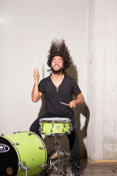 Mixed ethnicity millennial man in loft playing green drum set hair flying.
