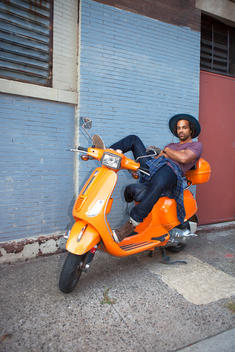 Outdoor street portrait of hipster athletic black male model in urban clothing sitting on an orange Vespa scooter wearing a hat