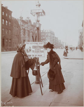 A Woman From The Salvation Army Standing Next To The Salvation Army Christmas Dinner Kettle. Another Woman Puts Money In The Pot. The Sign Above The Pot Says 
