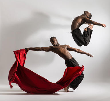 Two black male dancers jumping with red satin prop against white background